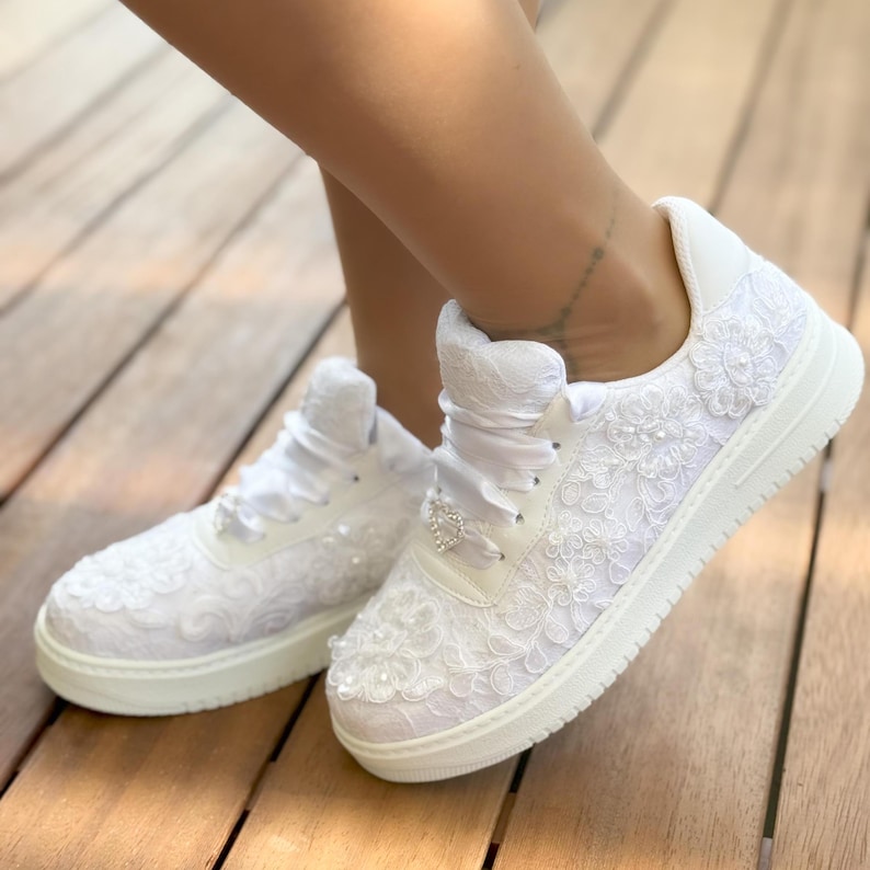 May include: White lace sneakers with a satin ribbon and a rhinestone heart detail. The sneakers have a white sole and are decorated with white lace.