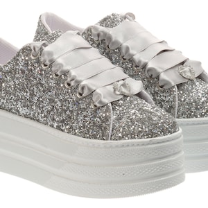 Puede incluir: Zapatillas de plataforma de glitter plateado con suela de plataforma blanca y una cinta de satén atada alrededor del tobillo.