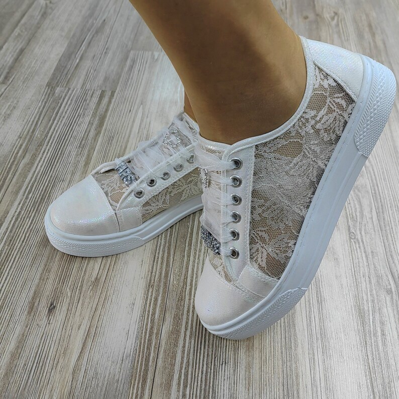 K&ouml;nnte beinhalten: Ein Paar wei&szlig;e Sneaker mit einem zarten floralen Spitzen&uuml;berzug an den Seitenteilen, einer schimmernden perlmuttfarbenen Zehenkappe und wei&szlig;en Band-Schn&uuml;rsenkeln, die mit silberfarbenen Strassverzierungen an den &Ouml;sen versehen sind.