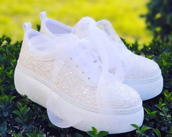 May include: White glitter sneakers with a white satin ribbon tied around the laces. The sneakers have a thick white platform sole.