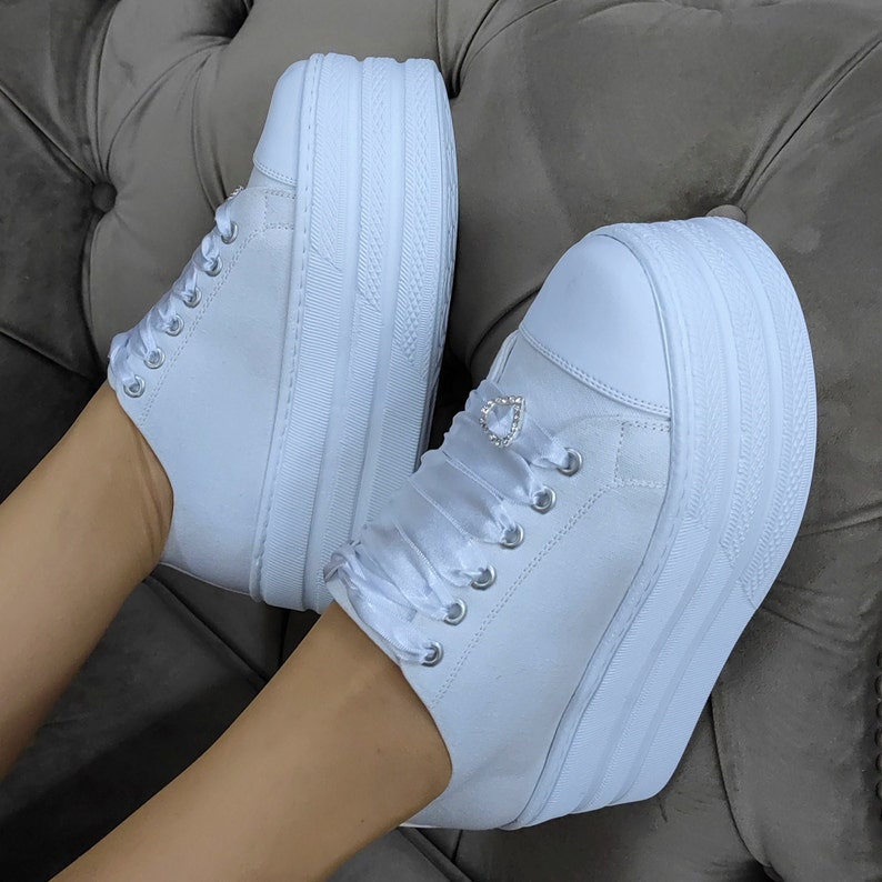 May include: A pair of white platform sneakers with a satin ribbon tied around the ankle. The sneakers have a heart-shaped rhinestone embellishment on the ribbon.