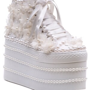 May include: White platform sneakers with a high top, embellished with pearl beads and white fabric flowers. The laces are white satin ribbon.