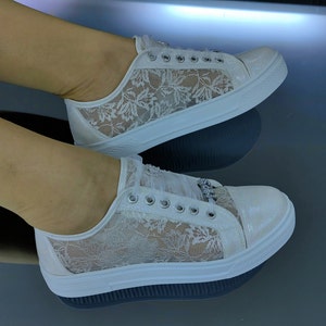 K&ouml;nnte beinhalten: Ein Paar wei&szlig;e Sneaker mit einem zarten floralen Spitzen&uuml;berzug an den Seitenteilen, einer schimmernden, irisierenden Zehenkappe, silberfarbenen &Ouml;sen und einem dekorativen, mit Kristallen verzierten Anh&auml;nger an den Schn&uuml;rsenkeln auf einer dicken Sohle.