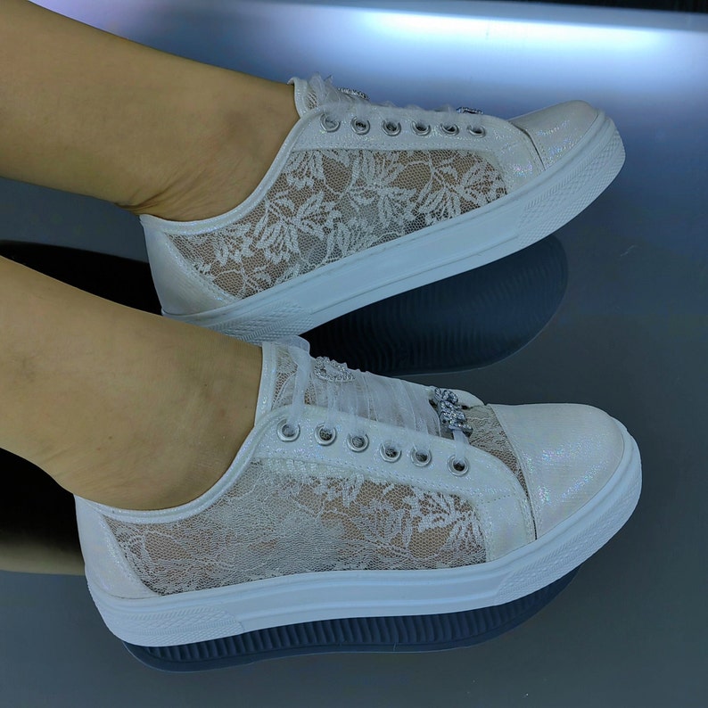 K&ouml;nnte beinhalten: Ein Paar wei&szlig;e Sneaker mit einem zarten floralen Spitzen&uuml;berzug an den Seitenteilen, einer schimmernden, irisierenden Zehenkappe, silberfarbenen &Ouml;sen und einem dekorativen, mit Kristallen verzierten Anh&auml;nger an den Schn&uuml;rsenkeln auf einer dicken Sohle.