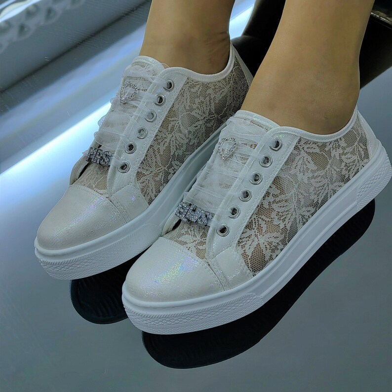 K&ouml;nnte beinhalten: Ein Paar wei&szlig;e Spitzen-Sneaker mit schimmernder, irisierender Zehenkappe, silberfarbenen &Ouml;sen und dekorativen Strassverzierungen an den Schn&uuml;rsenkeln, darunter ein herzf&ouml;rmiger Anh&auml;nger und ein rechteckiges Element mit klaren Kristallen.