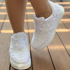 May include: White lace sneakers with a heart-shaped rhinestone embellishment on each shoe. The sneakers have a white satin ribbon tied around the ankle.