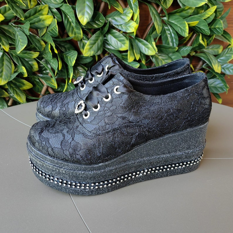 May include: Black lace platform shoes with a rhinestone trim. The shoes have a thick platform sole and a lace-up design.