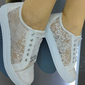 K&ouml;nnte beinhalten: Ein Paar wei&szlig;e Sneaker mit einem zarten floralen Spitzen&uuml;berzug an den Seitenteilen, einem schimmernden irisierenden Finish an Zehenkappe und Ferse, silberfarbenen Metall&ouml;sen, wei&szlig;en Band-Schn&uuml;rsenkeln und einer dicken wei&szlig;en Gummisohle mit dunklem Profil.