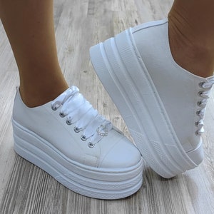 May include: White platform sneakers with a satin ribbon and a small heart-shaped charm. The sneakers have a thick platform sole.