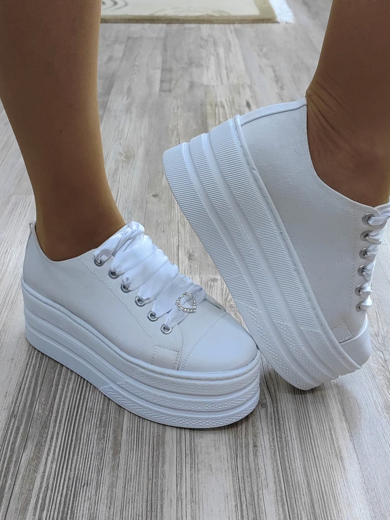 May include: White platform sneakers with a satin ribbon and a small heart-shaped charm. The sneakers have a thick platform sole.