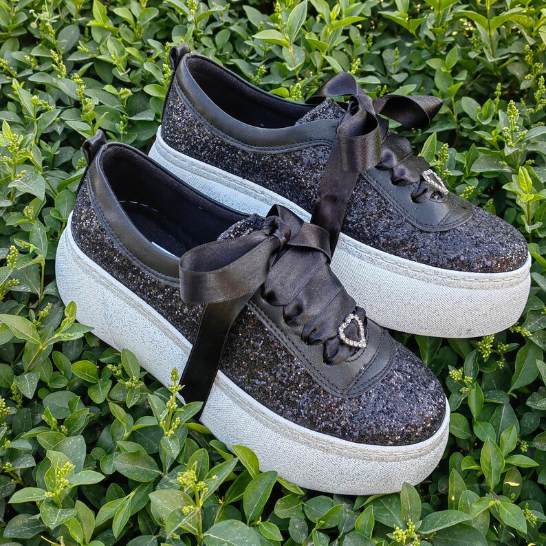 May include: Two pairs of black glitter sneakers with white soles and black satin ribbon laces. One pair has a heart-shaped rhinestone embellishment.