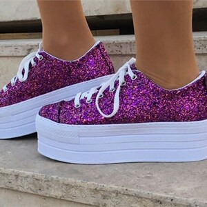 May include: A pair of platform sneakers with a vibrant purple glitter finish. The shoes have white laces and a thick white sole. The platform sole adds height to the shoes. The sneakers are a stylish footwear choice.
