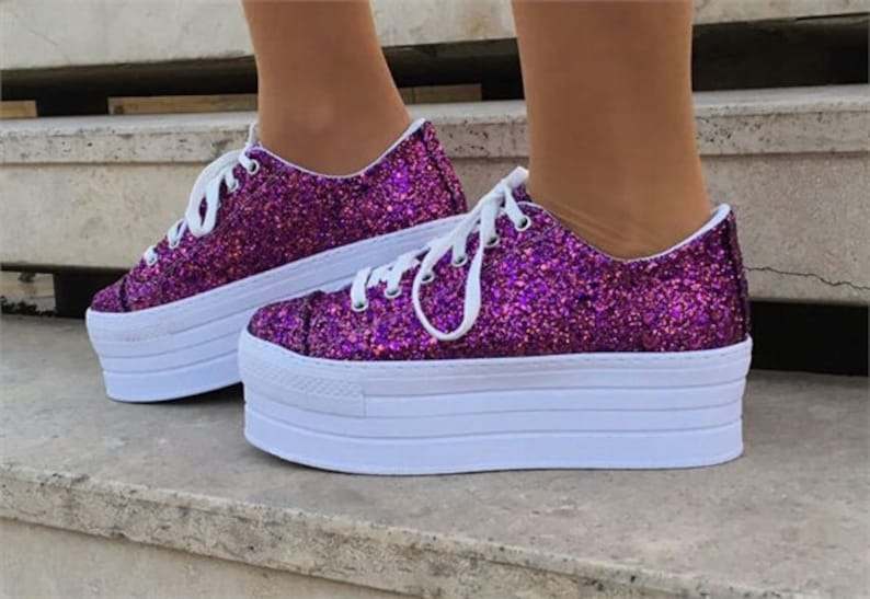 May include: A pair of platform sneakers with a vibrant purple glitter finish. The shoes have white laces and a thick white sole. The platform sole adds height to the shoes. The sneakers are a stylish footwear choice.