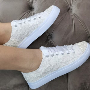Könnte beinhalten: Weiße Sneakers aus Spitze mit Perlenbesatz und Satinband-Schnürsenkeln. Die Schuhe haben eine weiße Gummisohle und sind perfekt für eine Hochzeit oder einen besonderen Anlass.