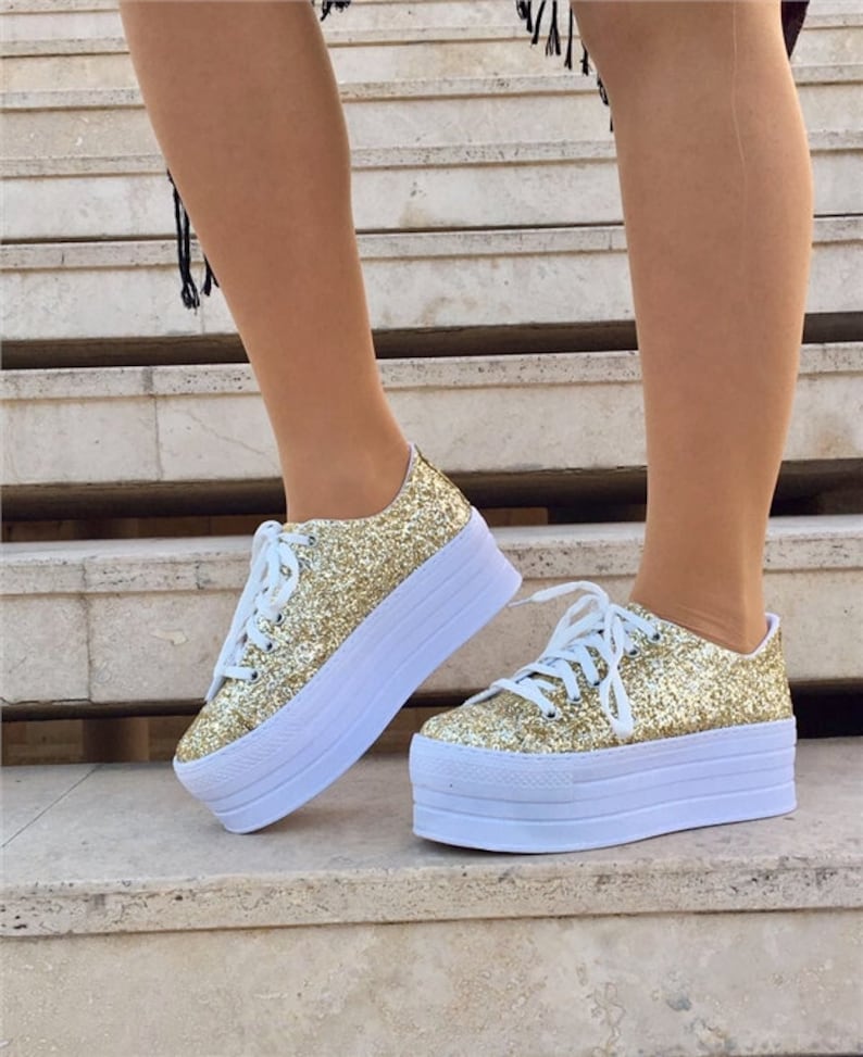 May include: Gold glitter platform sneakers with white laces and a thick white sole. The shoes have a sparkly, textured appearance and a classic lace-up design. The platform sole adds height and a modern touch to the footwear.