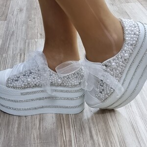 May include: White platform sneakers with a pearl and rhinestone embellishment. The sneakers have a white ribbon tied around the ankle.