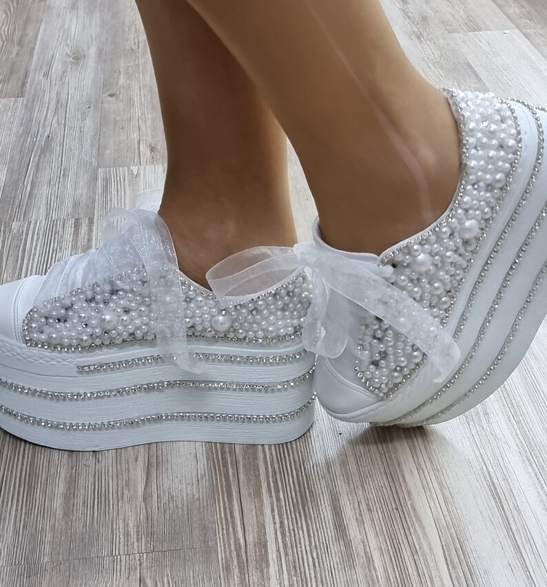 May include: White platform sneakers with a pearl and rhinestone embellishment. The sneakers have a white ribbon tied around the ankle.