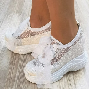 May include: White lace sneakers with a large white bow on the front. The sneakers have a thick white platform sole.