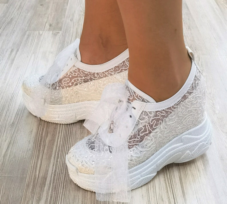 May include: White lace sneakers with a large white bow on the front. The sneakers have a thick white platform sole.