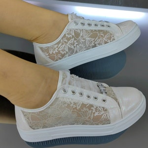 K&ouml;nnte beinhalten: Ein Paar wei&szlig;e Spitzensneaker mit schimmerndem, irisierendem Finish an den Zehen- und Fersenpartien. Die Seiten bestehen aus einem zarten wei&szlig;en Spitzen&uuml;berzug mit Blumenmuster. Die Schuhe verf&uuml;gen &uuml;ber silberfarbene &Ouml;sen und dekorative Kristallverzierungen.