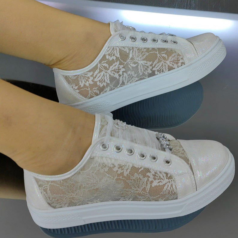 K&ouml;nnte beinhalten: Ein Paar wei&szlig;e Spitzensneaker mit schimmerndem, irisierendem Finish an den Zehen- und Fersenpartien. Die Seiten bestehen aus einem zarten wei&szlig;en Spitzen&uuml;berzug mit Blumenmuster. Die Schuhe verf&uuml;gen &uuml;ber silberfarbene &Ouml;sen und dekorative Kristallverzierungen.