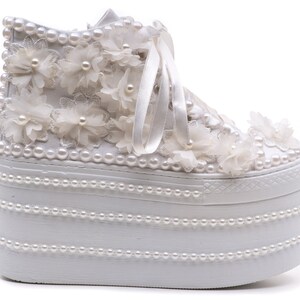 May include: White platform sneakers with a white lace and pearl embellishment. The sneakers have a thick platform sole and are decorated with white flowers and pearls.