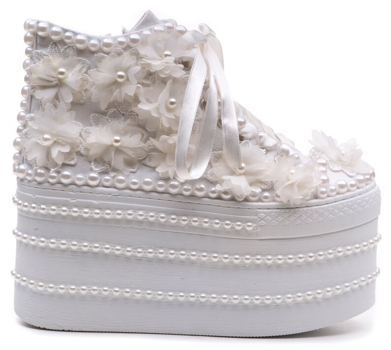 May include: White platform sneakers with a white lace and pearl embellishment. The sneakers have a thick platform sole and are decorated with white flowers and pearls.