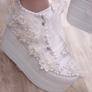 May include: White platform sneakers with a lace-up design and pearl embellishments. The shoes are decorated with white flowers and a heart-shaped rhinestone.