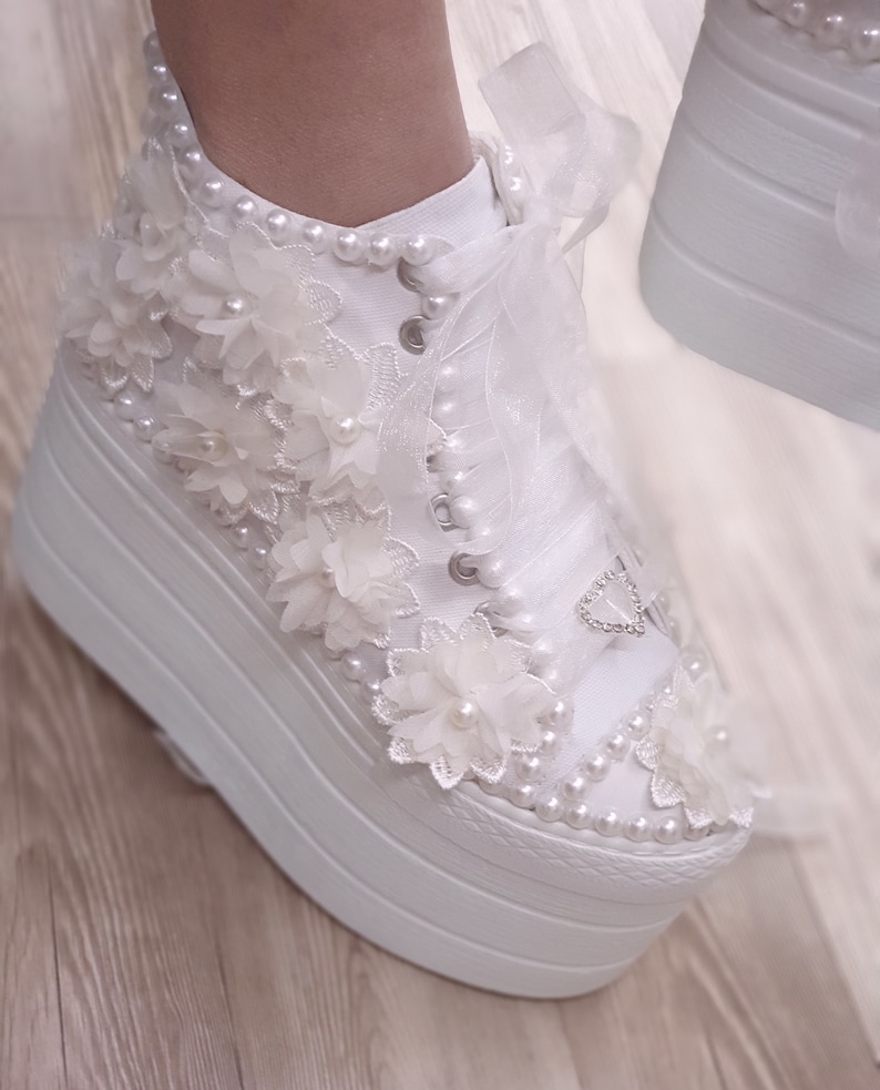 May include: White platform sneakers with a lace-up design and pearl embellishments. The shoes are decorated with white flowers and a heart-shaped rhinestone.
