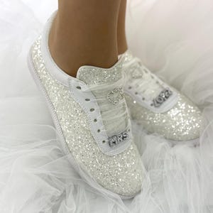 Può includere: Sneakers bianche glitterate con dettagli in pelle bianca. Le scarpe da ginnastica presentano un ciondolo a forma di cuore sui lacci e la scritta "MRS" sulla punta. I tacchi hanno la scritta "LOVE". Le scarpe sono su uno sfondo di tulle bianco.