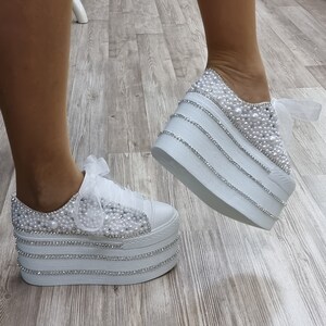 May include: White platform sneakers with a pearl and rhinestone embellishment. The sneakers have a white satin ribbon tied around the ankle.