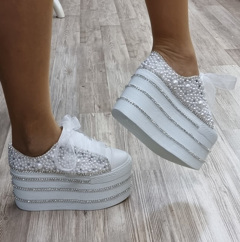 May include: White platform sneakers with a pearl and rhinestone embellishment. The sneakers have a white satin ribbon tied around the ankle.