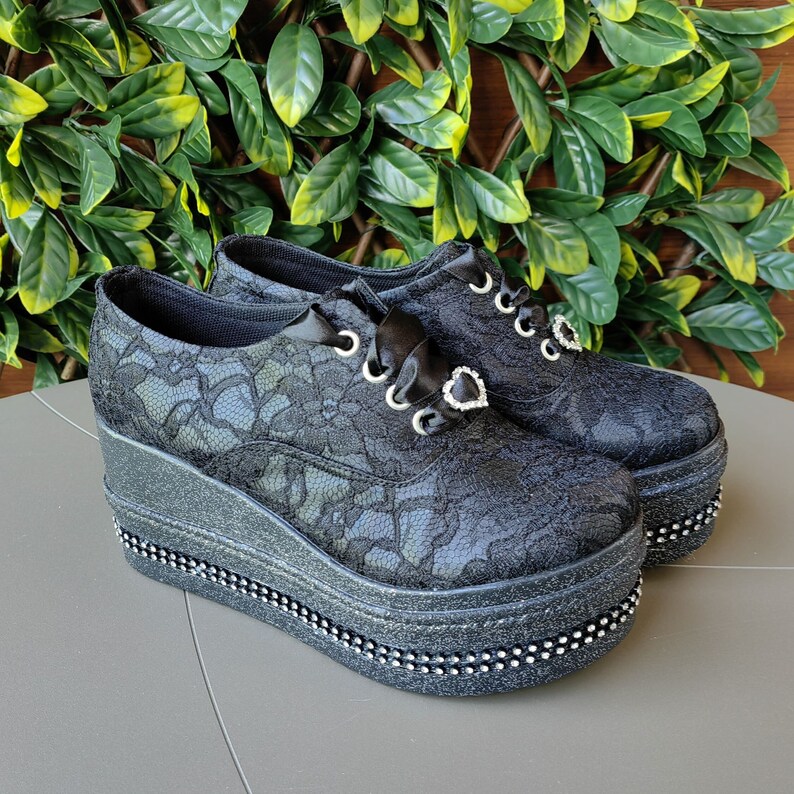 May include: Black lace platform shoes with a black ribbon and a rhinestone heart detail. The shoes have a thick platform sole with a beaded trim.