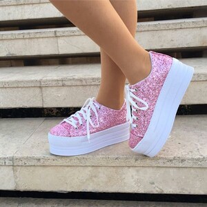 May include: Pink glitter platform sneakers with white laces and a white sole. The shoes are on a set of stone steps. The sneakers have a classic lace-up design and a thick, layered sole.