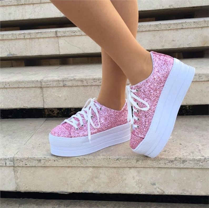 May include: Pink glitter platform sneakers with white laces and a white sole. The shoes are on a set of stone steps. The sneakers have a classic lace-up design and a thick, layered sole.