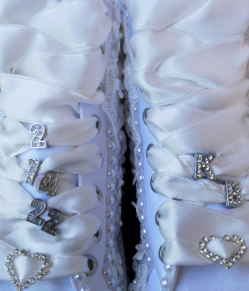 May include: A pair of white canvas shoes with white satin ribbon laces. The laces are tied with silver charms in the shape of numbers and a heart. The shoes are decorated with white pearl beads.
