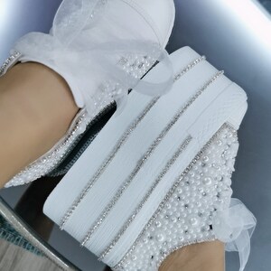 May include: White platform sneakers with a pearl and rhinestone design. The sneakers have a white satin ribbon tied around the ankle.