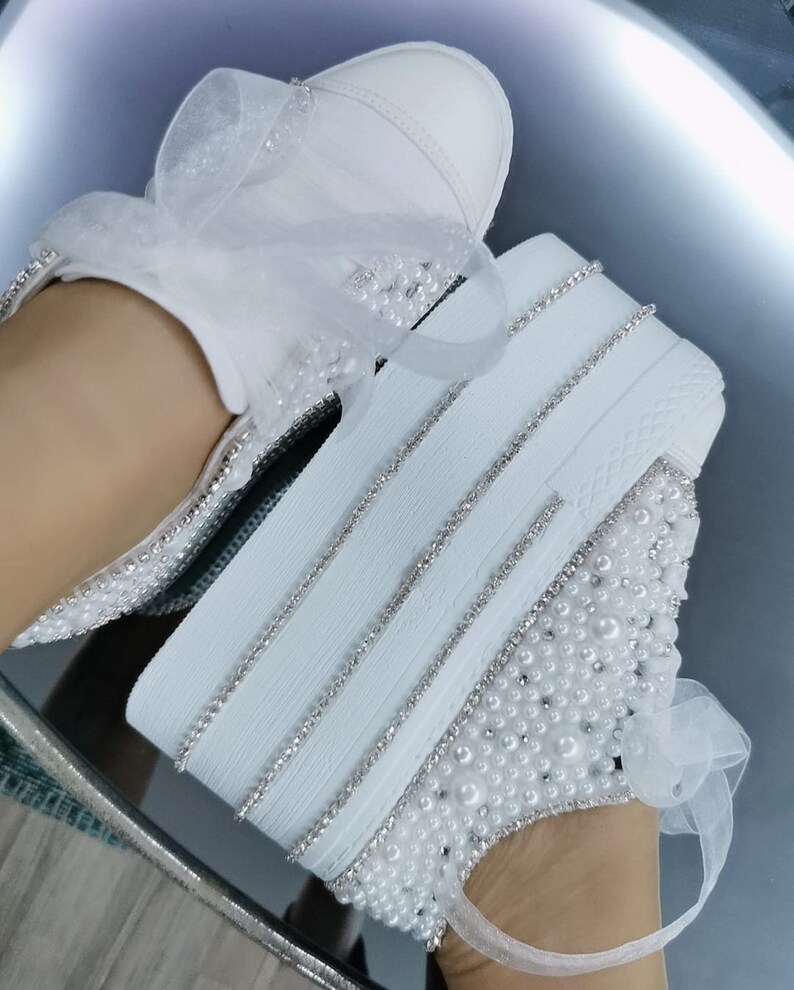 May include: White platform sneakers with a pearl and rhinestone design. The sneakers have a white satin ribbon tied around the ankle.