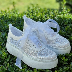 May include: White lace sneakers with a platform sole and a white ribbon tied around the ankle. The sneakers are decorated with sequins and a small rhinestone heart.