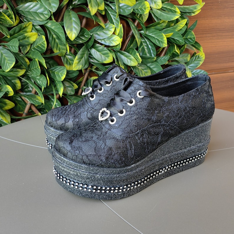 May include: Black platform shoes with a lace-up design and a decorative heart detail. The shoes have a chunky platform sole and a glitter finish.