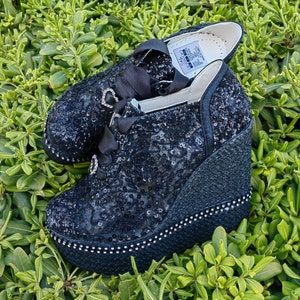 May include: A pair of black wedge shoes with a lace-up design and a sparkly finish. The shoes have a platform sole and a pointed toe.