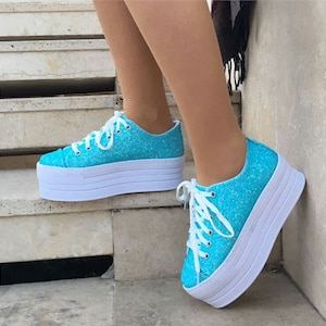 May include: Turquoise glitter platform sneakers with white laces and a thick white sole. The shoes are a vibrant blue and have a sparkly texture. The platform sole adds height and a modern touch to the footwear.