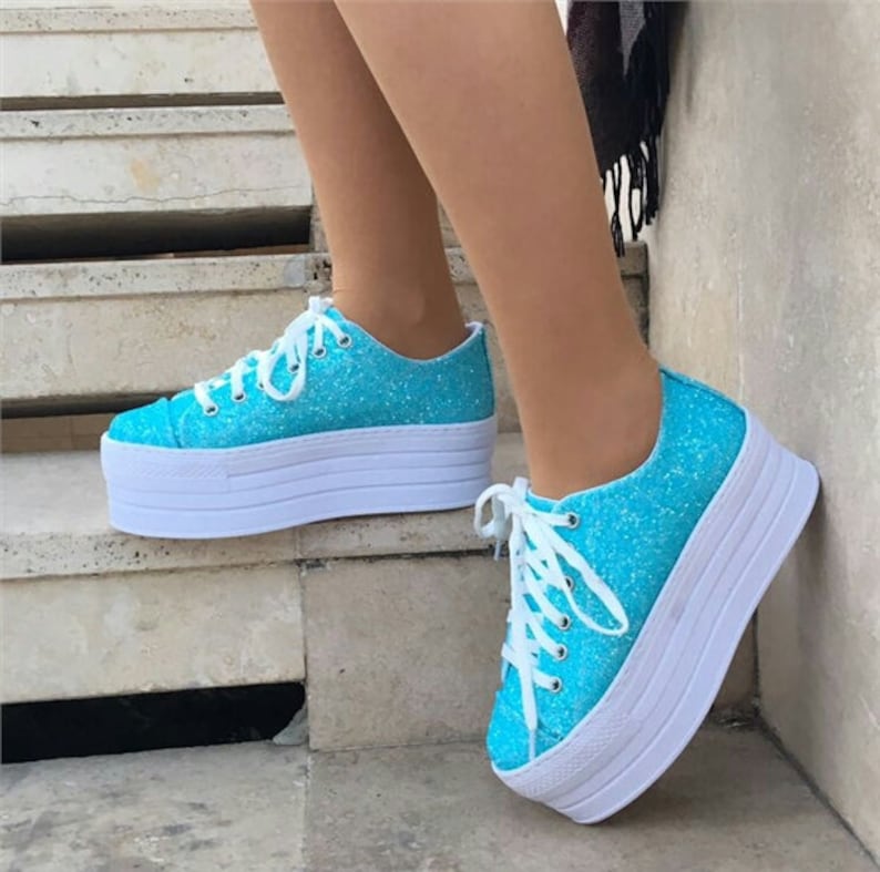 May include: Turquoise glitter platform sneakers with white laces and a thick white sole. The shoes are a vibrant blue and have a sparkly texture. The platform sole adds height and a modern touch to the footwear.