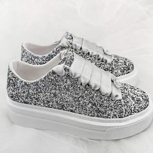 Puede incluir: Zapatillas de plataforma plateadas con purpurina y un lazo de satén atado alrededor de los cordones. Las zapatillas tienen una suela de plataforma blanca.
