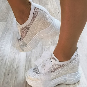 May include: White lace sneakers with a platform sole and a white tulle bow. The sneakers are decorated with rhinestones.
