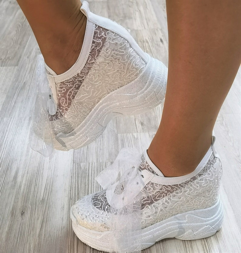 May include: White lace sneakers with a platform sole and a white tulle bow. The sneakers are decorated with rhinestones.