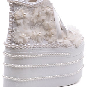 May include: White platform sneakers with a high platform sole decorated with pearl beads and white fabric flowers.