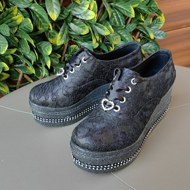 May include: Black lace platform shoes with a heart-shaped charm on the laces. The shoes have a thick platform sole and a black glitter trim.