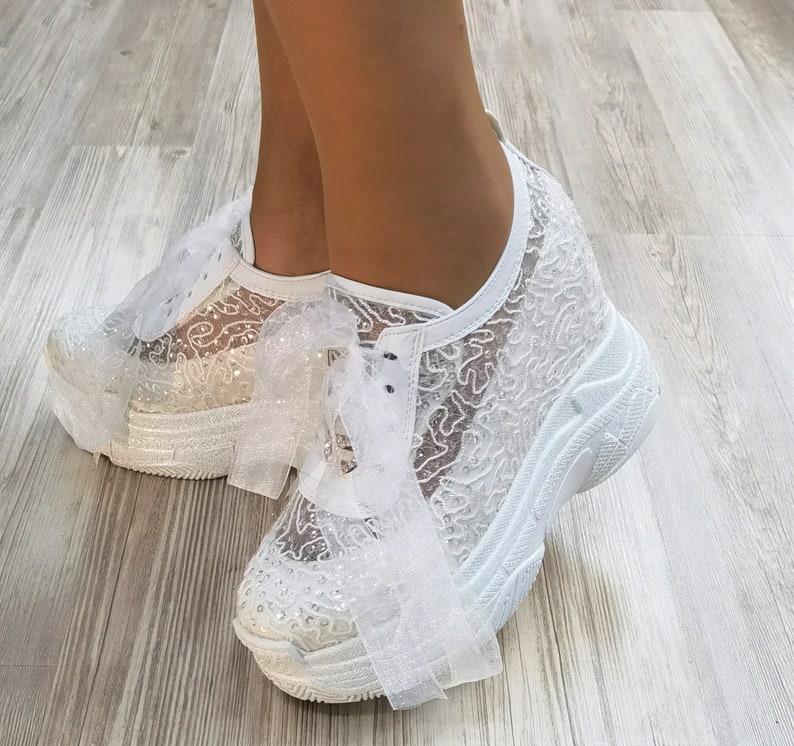 May include: White lace sneakers with a platform sole and a white ribbon bow. The sneakers are decorated with glitter.