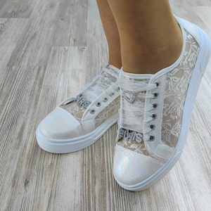 K&ouml;nnte beinhalten: Ein Paar wei&szlig;e Sneaker mit floralem Spitzen&uuml;berzug an den Seiten, schillernden Zehenkappen und transparenten Schleifenb&auml;ndern. Jeder Schuh ist mit einem silbernen herzf&ouml;rmigen Anh&auml;nger und einem silbernen Strass-Anh&auml;nger mit dem Wort ELVIS verziert.
